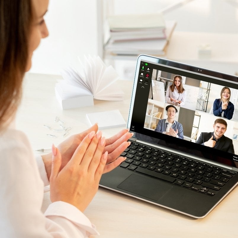 A woman sits at a desk clapping while participating in a Project Management for Remote Teams video call. Four people on her laptop screen are smiling and clapping, creating an engaging virtual meeting atmosphere.