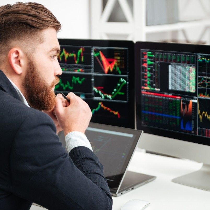 A man in a suit sits at a desk, closely analyzing stock charts and graphs alongside Project Progress Tracking in BVDash displayed on multiple computer monitors in an office setting.