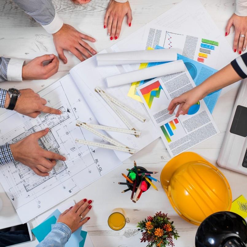 Four people collaborate at a table with blueprints, charts, documents, a yellow construction helmet, a laptop, and drawing tools, suggesting an agile project plan or architectural meeting in progress.