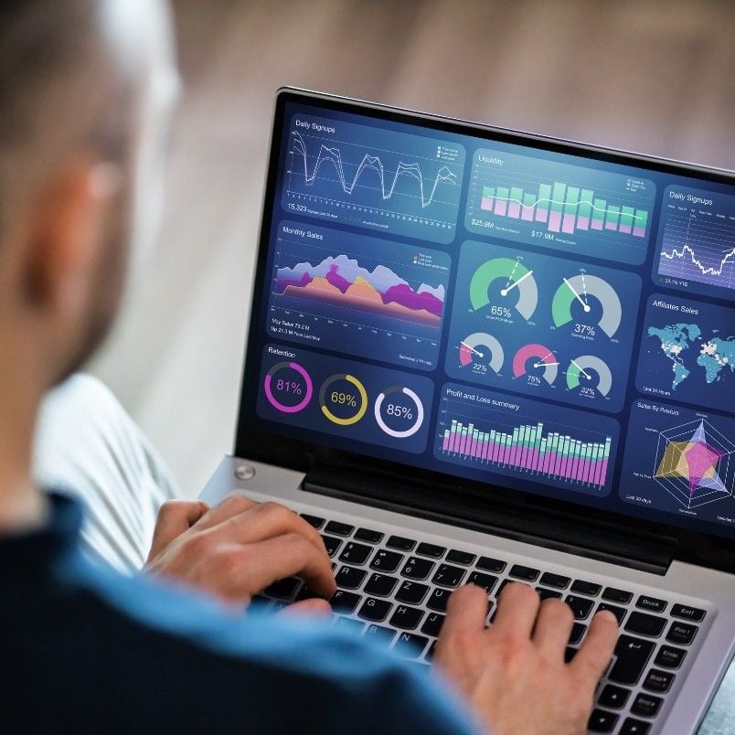 A person uses a laptop displaying colorful data dashboards and graphs, including pie charts, bar graphs, line charts, and Gantt Charts in BVDash, representing various analytics and project statistics.