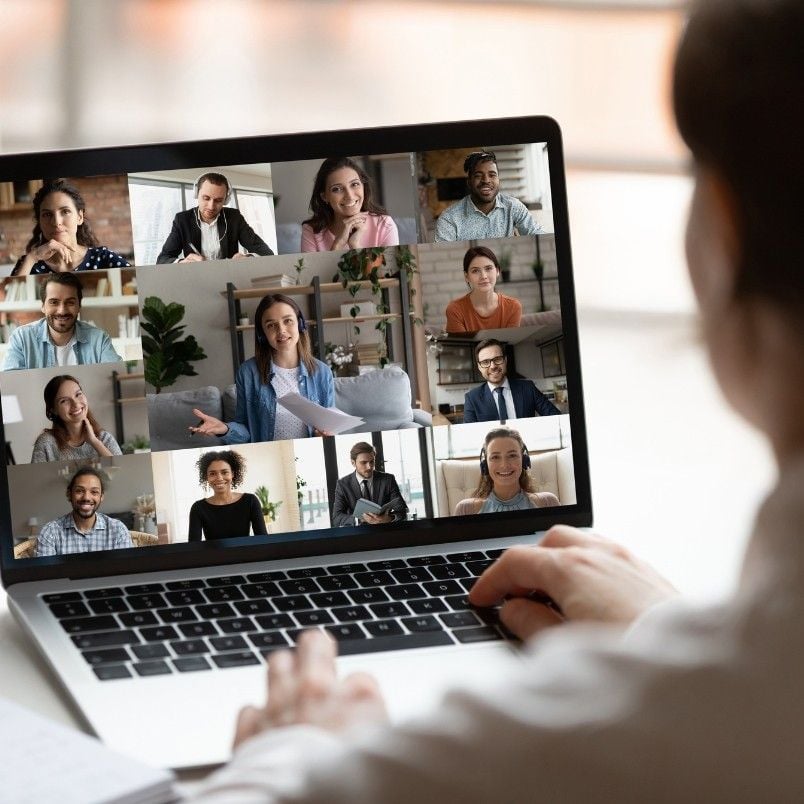 A person participates in a video conference on a laptop using BVDash for Stakeholder Communication, with twelve diverse people visible in separate windows, all smiling and appearing engaged.