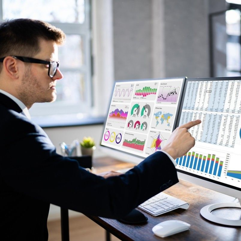 A man in a suit and glasses analyzes charts and data on dual computer monitors at a modern desk, pointing to graphs and spreadsheets as he conducts risk monitoring in BVDash.