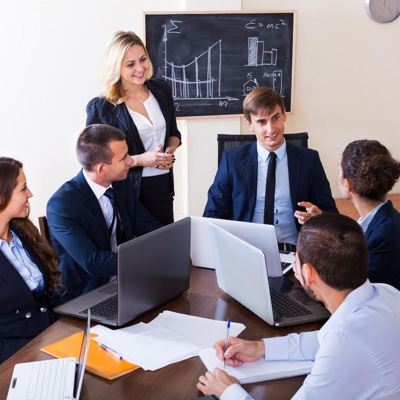 Six business professionals in suits have a meeting around a table with laptops and papers. A woman stands smiling while the others discuss Example PMP® Questions. A chalkboard with graphs and formulas is visible in the background.