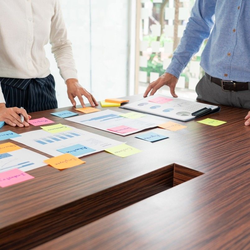 Two people in business attire stand at a wooden table covered with charts, graphs, and colorful sticky notes, reviewing documents and data as they collaborate on an agile project plan during a meeting. Only their torsos and hands are visible.