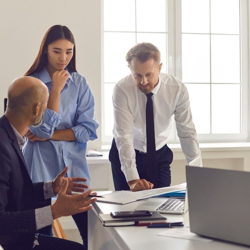 Three colleagues in business attire are having a serious discussion around a desk with papers, a laptop, and notes on Initiative vs Epic, in a bright office with large windows. One man gestures while speaking as the others listen attentively.