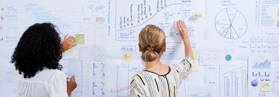 Two people stand before a wall covered with charts, graphs, and diagrams, organizing Epics, Features, and User Stories using sticky notes and markers as they collaborate on project planning.