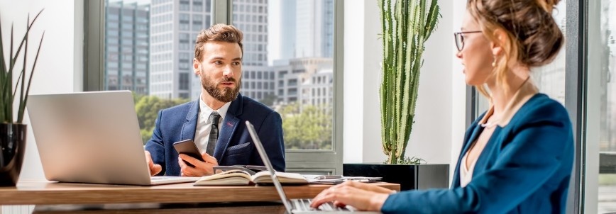 Two business professionals in formal attire sit at a desk in a modern office. The man discusses Example PMP® Questions on his smartphone while the woman types on a laptop. Tall plants and city buildings are visible through large windows in the background.