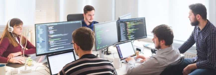 Five people work together in an office, seated at desks with multiple monitors displaying code. One person wears headphones; others discuss software development management, collaborating on programming tasks.