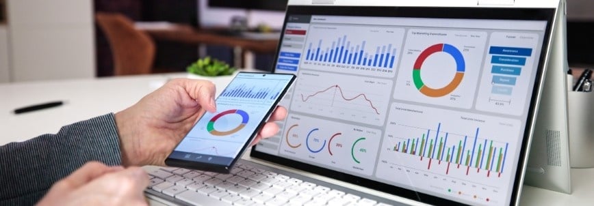 A person holds a smartphone while viewing a laptop screen displaying colorful charts and graphs on the BVDash Dashboard, including pie charts, bar graphs, and line graphs, in a modern office setting.