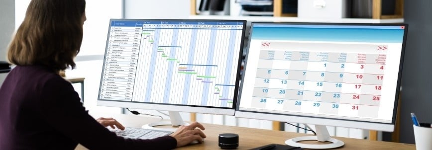 A person sits at a desk using two computer monitors; one screen displays KPI Tracking in BVDash alongside a Gantt chart, while the other shows a monthly calendar.