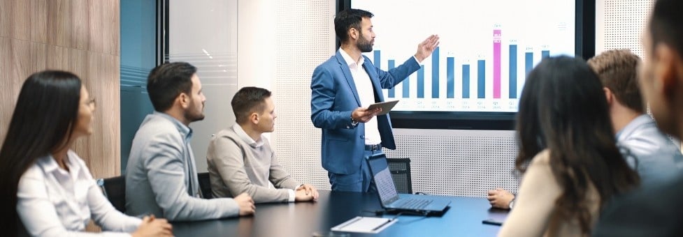 A man in a suit presents data on Task Management in BVDash to five colleagues seated around a conference table in a modern office meeting room. The screen displays a colorful bar chart.