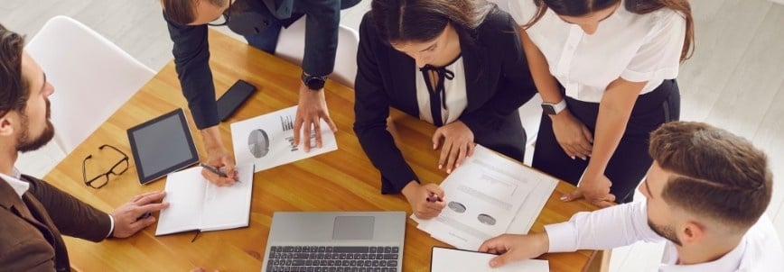 Five business professionals gather around a wooden table, reviewing documents and charts, discussing the differences between an initiative vs epic as they collaborate on a project in a bright office setting.