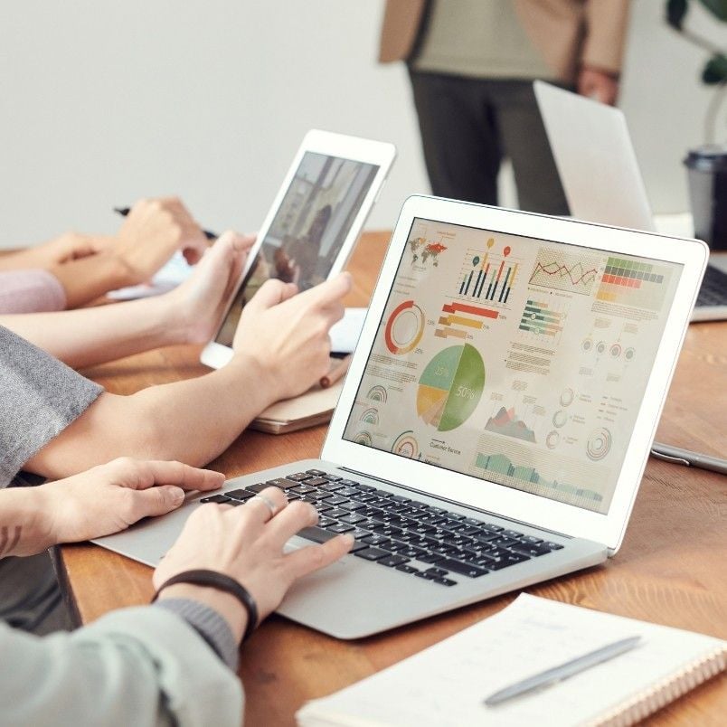People sit at a table using laptops and tablets. One laptop screen displays colorful charts and Gantt Charts in BVDash. A notebook, pen, and a blurred person standing in the background are also visible.