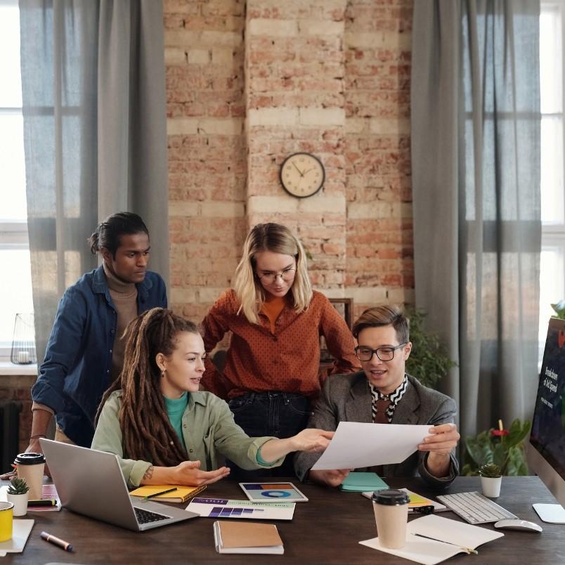 Four people gather around a table with laptops and documents, discussing an agile project plan one person is holding, in a modern office with a brick wall, large windows, and a clock on the wall.