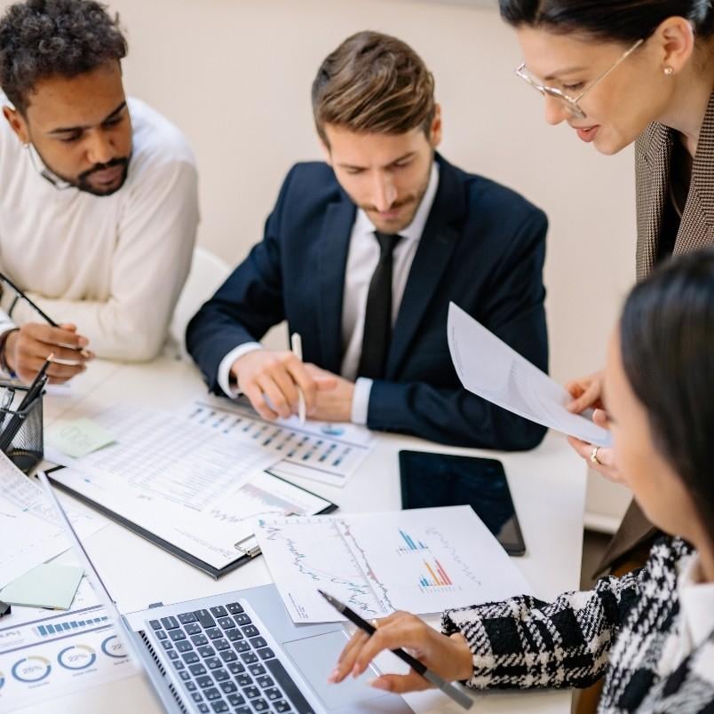 Four business professionals are gathered around a desk with documents, charts, a laptop, and a tablet, discussing financial graphs and data while debating Initiative vs Epic during a meeting.