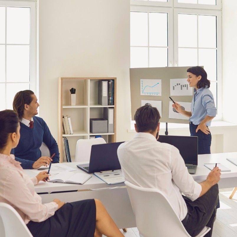 A woman stands by a flip chart with graphs, explaining Initiative vs Epic to three colleagues seated at a table with laptops and documents in a bright office.