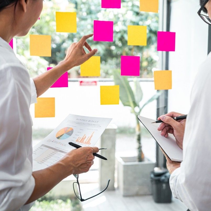Two people stand near a window covered with colorful sticky notes, discussing project deadlines in BVDash. One points to a note while the other writes in a notebook, and a report with charts and graphs is held. Sunlight and greenery shine outside.