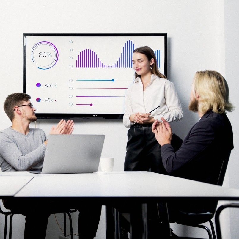 Three people are in a meeting room. A woman presents data on risk monitoring in BVDash on a large screen, while two men sit at the table with a laptop, applauding the colorful charts and percentage statistics displayed.