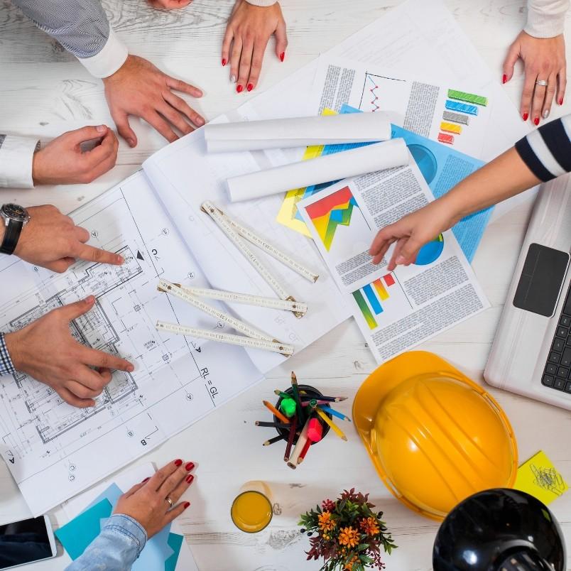 Several people collaborate around a table with blueprints, charts, documents, a laptop, ruler, yellow hard hat, and a small plant—blending architectural planning with Software Development Management in a dynamic team meeting.