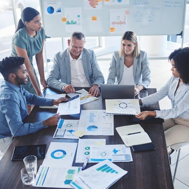 Five professionals sit around a table covered with charts and documents, discussing software development management. A whiteboard with graphs is visible in the background, while one person uses a laptop and others review printed reports.
