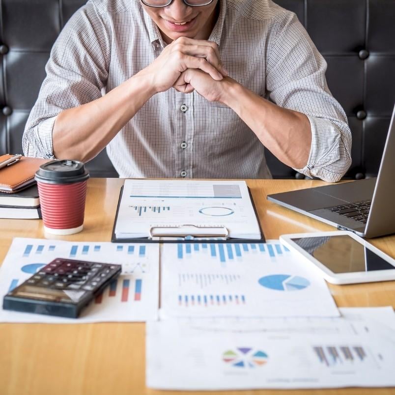 A person sits at a desk with financial charts, graphs, a tablet, a laptop, and documents, appearing focused as they review business data and analyze reports related to software development management.