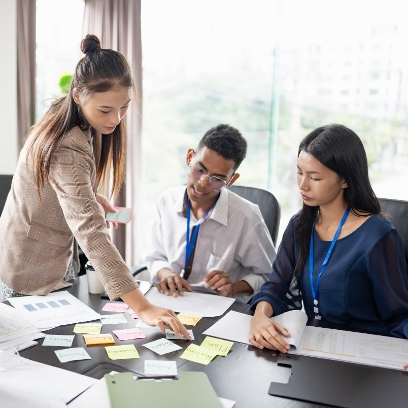 Three people sit at a table covered in papers and sticky notes, collaborating on an agile project plan. One person stands, pointing at the notes, while the others listen and take notes. Large windows are in the background.
