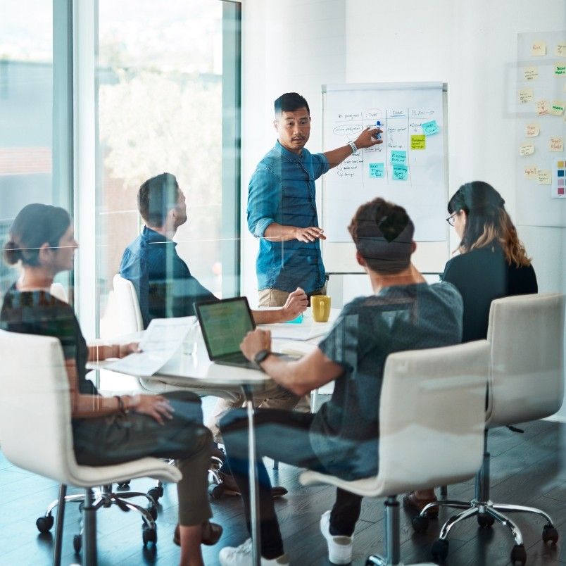 A man stands by a whiteboard with sticky notes, leading a discussion with four colleagues around a table in a modern office meeting room, as Example PMP® Questions appear on laptops and documents scattered across the table.