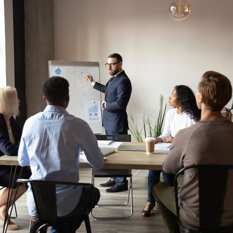 A man in a suit stands next to a whiteboard giving a presentation on Project Deadlines in BVDash to four colleagues seated around a table, with charts and notebooks in front of them, in a modern office setting.