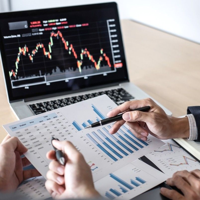 Two people discuss financial charts and graphs on printed sheets, pointing with pens. In the background, a laptop displays a candlestick stock chart, highlighting effective risk monitoring in BVDash amidst fluctuating market values.