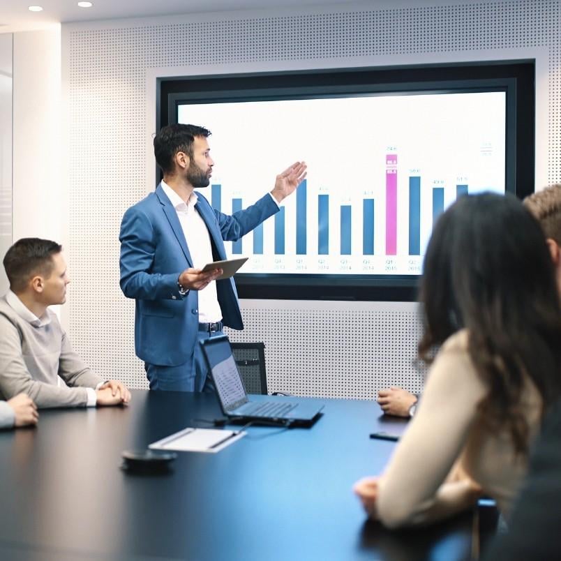 A man in a blue suit presents a bar graph on a large screen to four colleagues seated around a conference table, illustrating a Project Risk Assessment in BVDash during a business meeting or data presentation.
