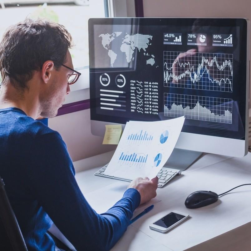 A person sits at a desk analyzing charts and graphs on printed sheets and a computer monitor displaying data visualizations, including world maps, line graphs, and Gantt Charts in BVDash, in a modern office setting.