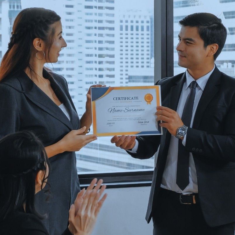 Two people in business attire stand indoors; one is handing a certificate for completing Example PMP® Questions to the other, while a third person applauds in the foreground. Large windows reveal tall buildings in the background.
