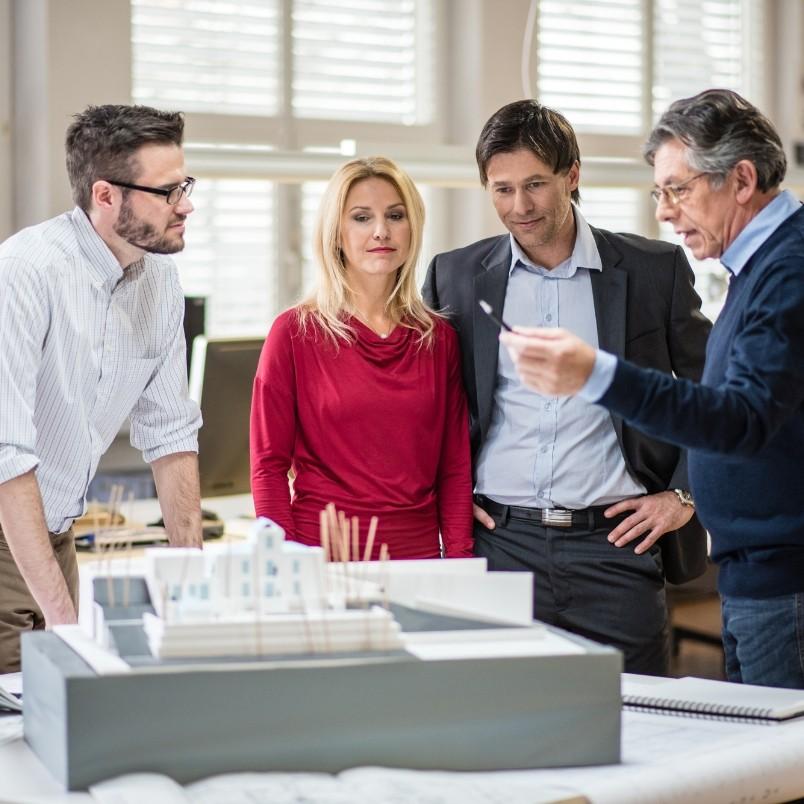 Four people stand around an architectural model in an office, discussing design plans and referencing example PMP® questions. One man gestures with a pen while the others listen attentively. Blueprints and models are on the table.