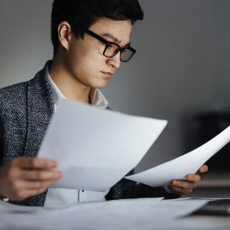 A person wearing glasses and a suit jacket is holding and reading multiple sheets of paper at a desk, appearing focused as they review a Project Risk Assessment in BVDash.