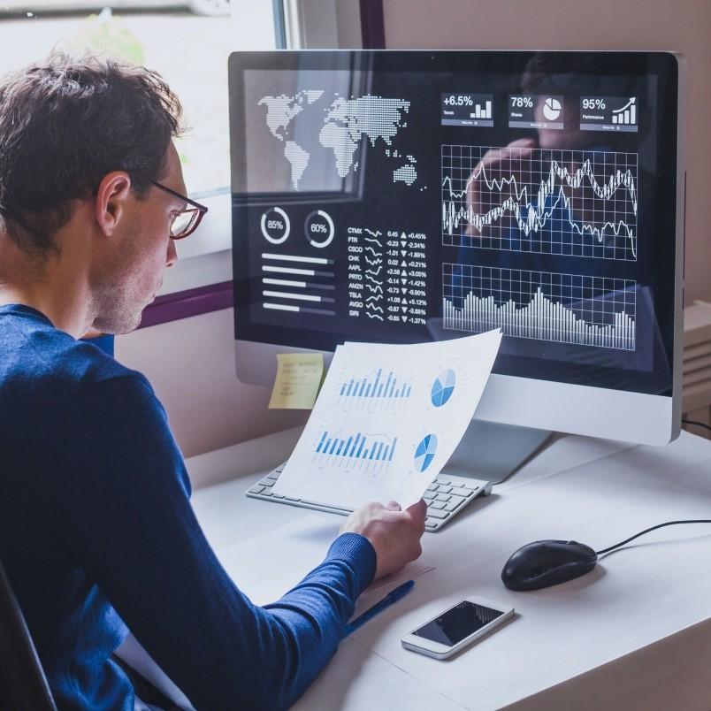 A man sits at a desk analyzing printed charts while looking at data visualizations, graphs, and world maps for risk monitoring in BVDash on his computer monitor. A smartphone, mouse, and keyboard are placed on the desk.