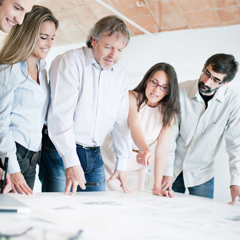 Five people stand around a table, closely examining documents together as they discuss the scope of an initiative vs epic. They appear engaged and focused, collaborating on a project in a bright, modern workspace.