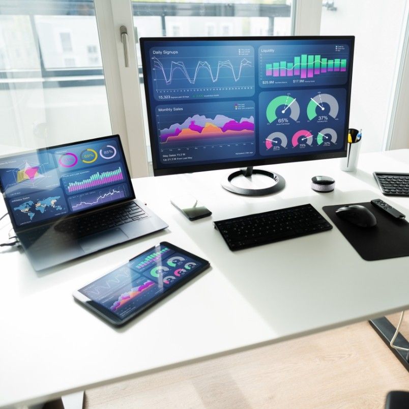 A modern desk setup with a monitor, laptop, and tablet displaying colorful charts, graphs, and Notion Progress Bars for data analysis. A keyboard, mouse, phone, notebook, and pens are neatly arranged on the white desk.