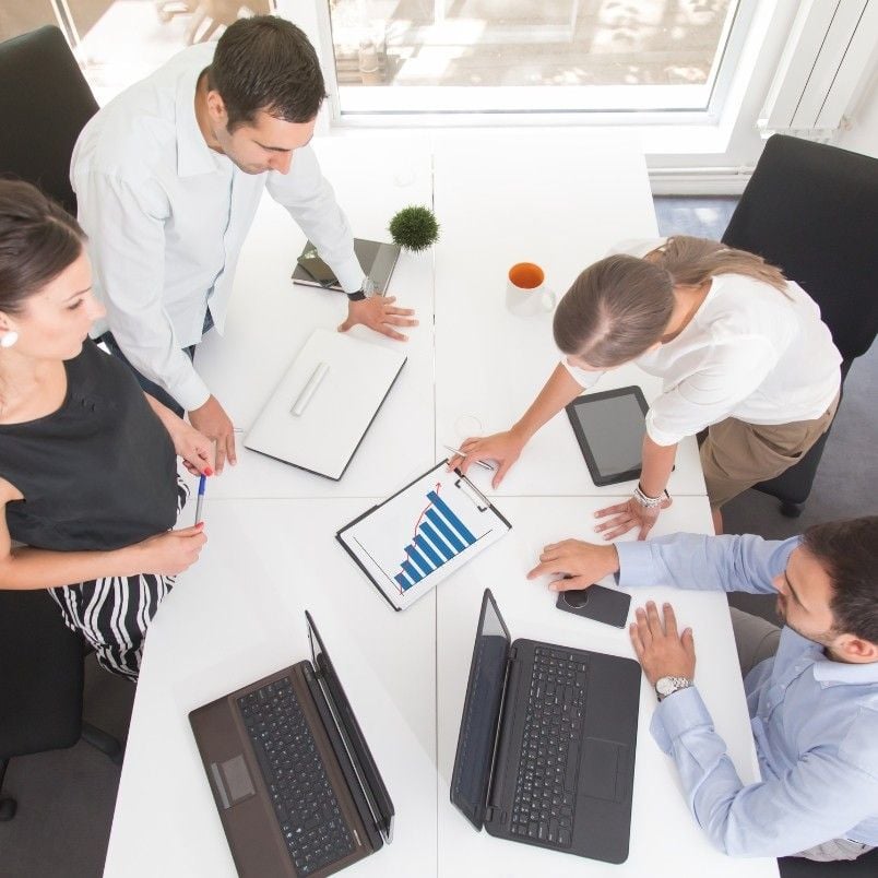Four people gathered around a white table with laptops and tablets, engaged in Sprint Planning as they discuss a bar graph displayed on a tablet during their collaborative business meeting.