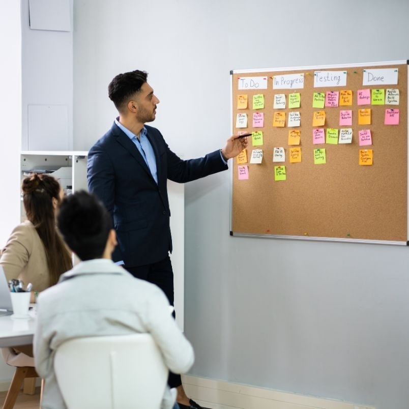 A man in a suit points to colorful sticky notes arranged in columns on a corkboard, labeled "To Do," "In Progress," "Testing," and "Done," illustrating Agile Planning as two seated people listen attentively.