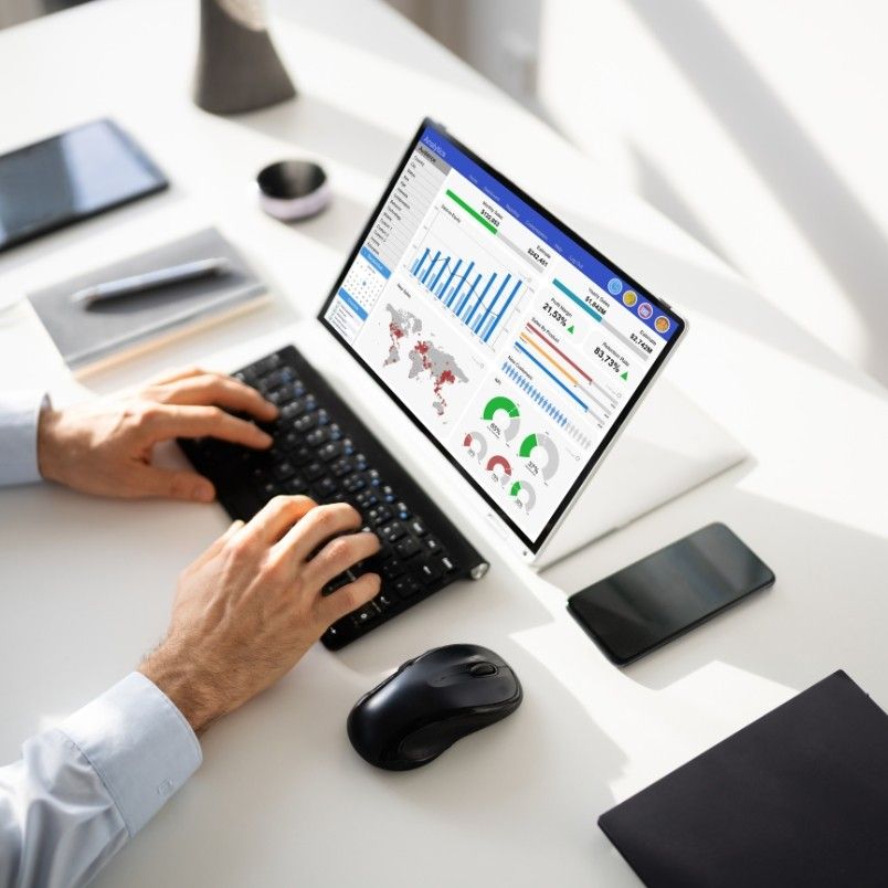 Person working at a desk with a laptop displaying data analytics dashboards, charts, and Notion Progress Bars. The workspace includes a keyboard, mouse, smartphone, notebook, and tablet in an organized office setting.