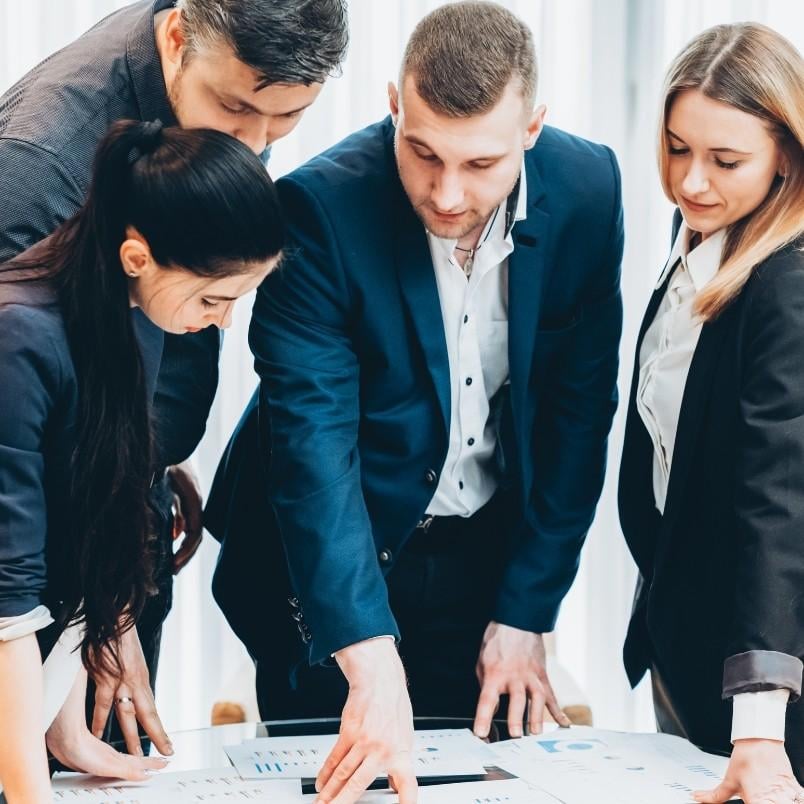 Four business professionals stand around a table, closely examining documents and charts together, suggesting collaborative sprint planning and teamwork in a modern office setting.