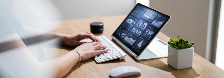 Person typing on a wireless keyboard at a wooden desk with a tablet displaying Data Analytics in BVDash and charts, next to a mouse, small speaker, and potted plant.