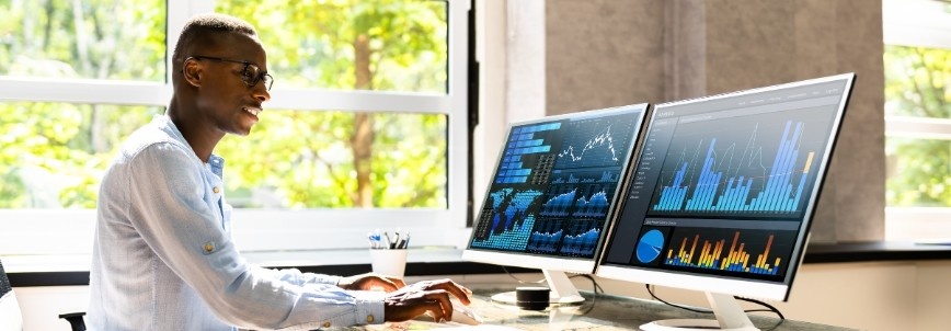 A person sits at a desk in front of two large monitors displaying colorful graphs and charts, managing Content Management in Notion, working in a bright office with sunlight streaming through a window.