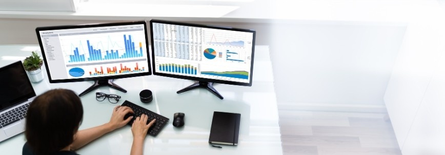 Person working at a desk with two monitors displaying charts and graphs, showcasing IoT Integration with BVDash, along with a keyboard, plant, notebook, and cup on a white surface in a bright office environment.