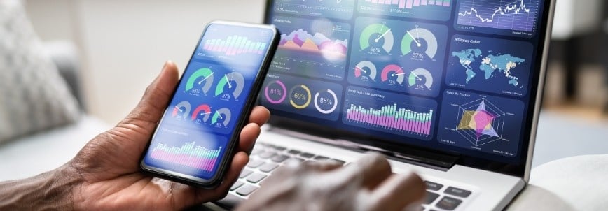 A person holds a smartphone displaying colorful charts and graphs, while a laptop nearby shows multiple dashboards with data visualizations in BVDash, including bar graphs, line charts, and a world map for managing files efficiently.
