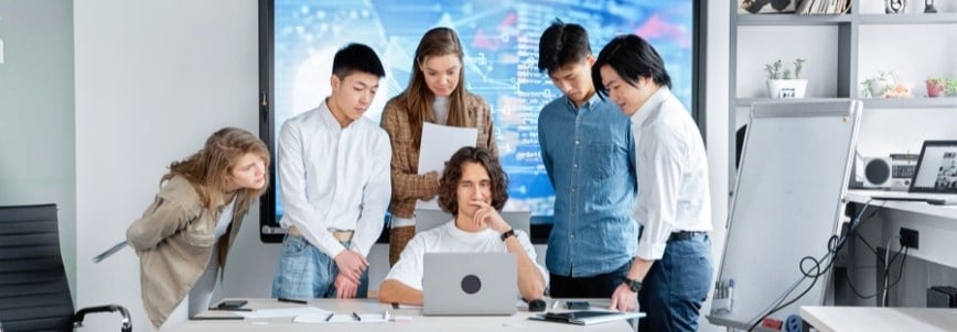 Six young adults gather around a desk, focused on a laptop. Charts and data about their Company Wiki in Notion are displayed on a large screen, suggesting collaborative discussion in a modern office setting.
