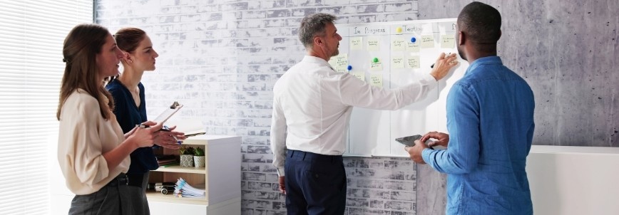 Four people in business attire stand around a whiteboard covered with sticky notes, actively engaged in Agile Planning. One writes on the board while others observe, take notes, or hold a tablet in a modern, well-lit office setting.
