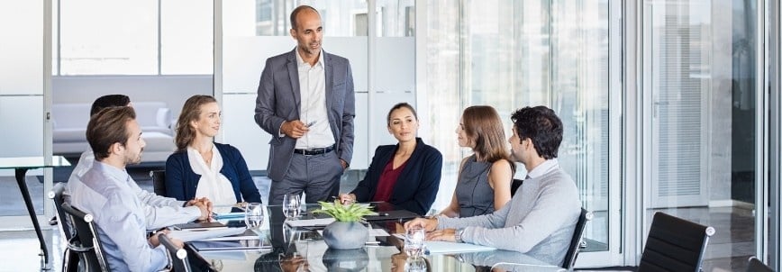 A group of business professionals sit around a conference table in a modern office, listening to a man in a suit discuss stakeholder support. Laptops, documents, and glasses of water are on the table.