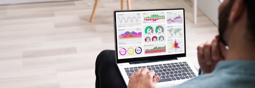 A person sits and looks at a laptop displaying colorful data visualizations, including graphs, charts, and maps, while exploring File Management in BVDash in a bright indoor setting.