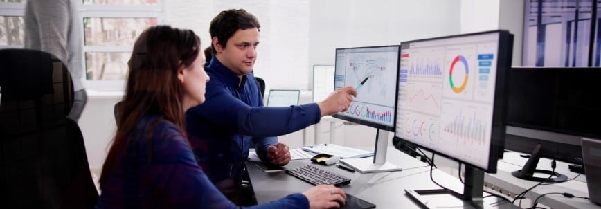 Two colleagues sit at a desk with multiple monitors displaying charts and graphs powered by Data Automation in BVDash. One points at data on a screen, while the other looks on attentively, highlighting their collaborative work environment.